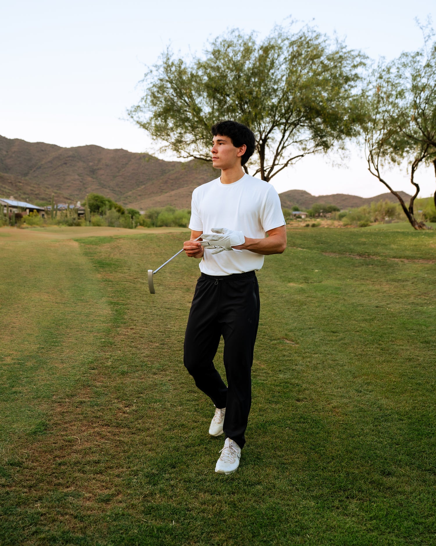 ALL IN ALL model is on the golf course green with his club and pulling off his golf gloves. He is wearing the off-white supernatural bamboo tee and black supernatural 2 joggers. 