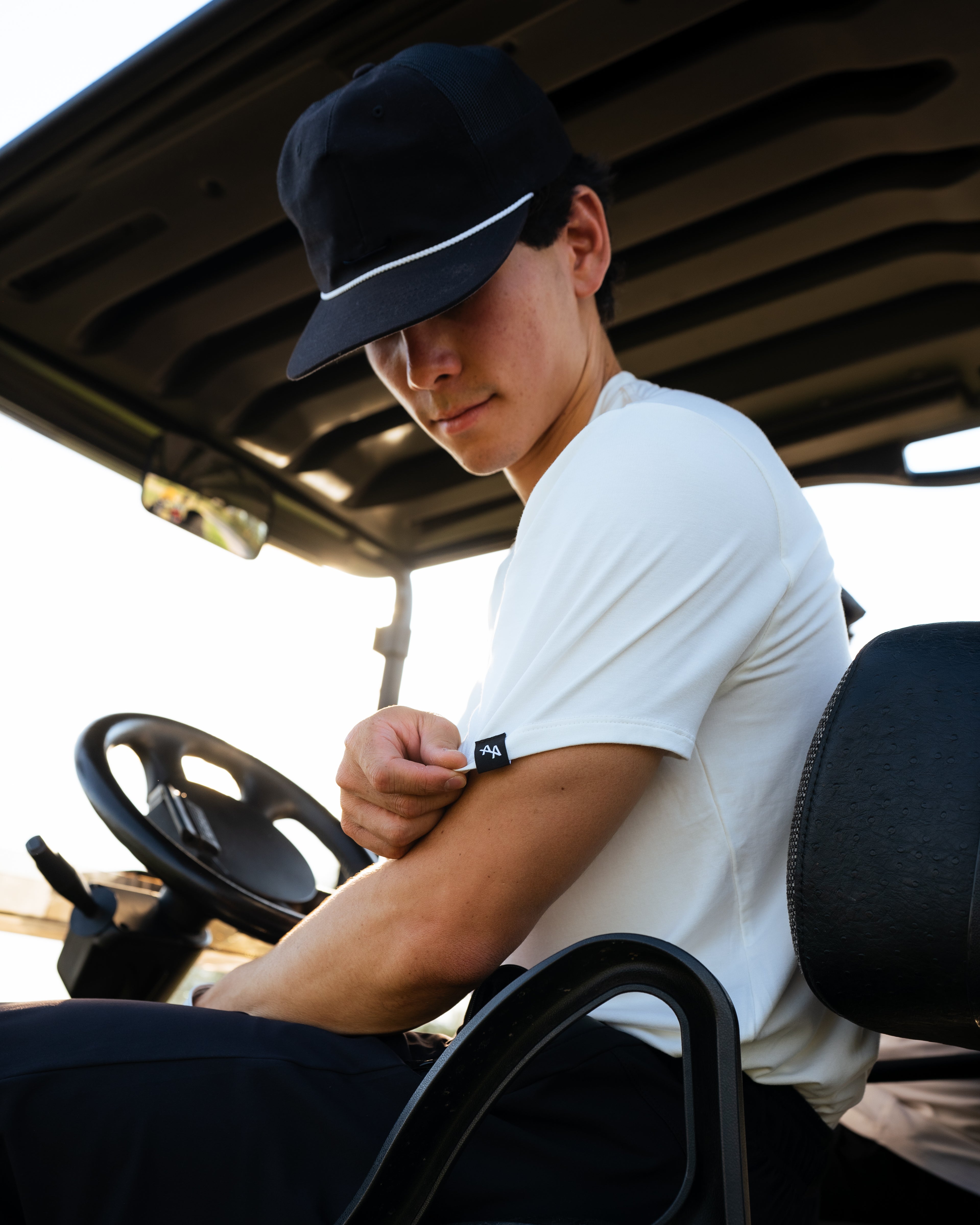 ALL IN ALL model is backlit and sitting in a golf cart. He is tugging on the left sleeve of an off-white bamboo tee highlighting the black and white double A logo.