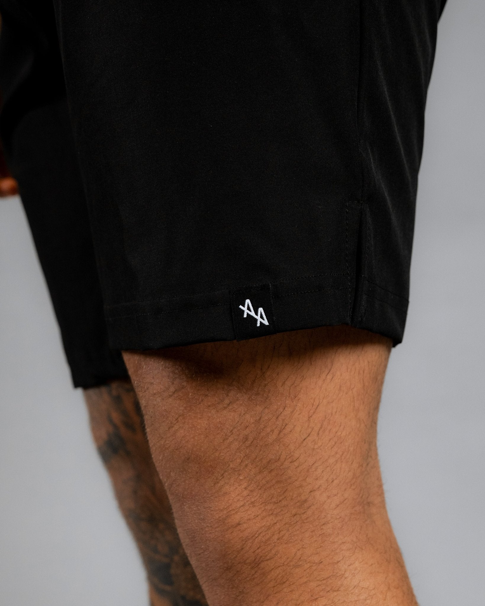 close up of woven flag label sewn onto the hem of the left leg of the black supernatural shorts. Flag features the white double "A" ALL IN ALL logo on a black woven tag.