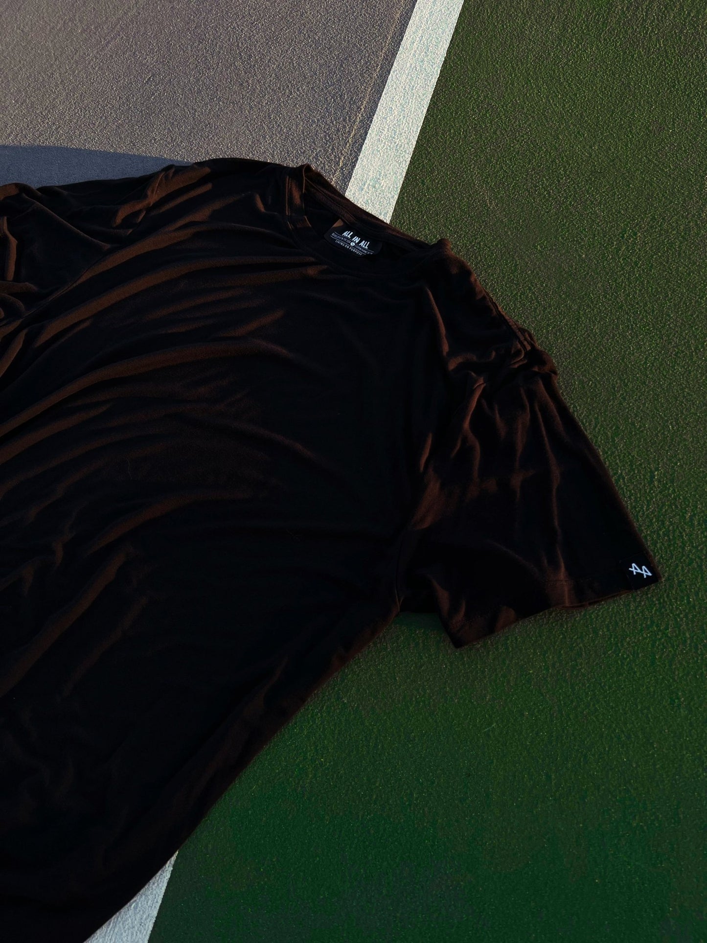 dramatic photo of ALL IN ALL black supernatural tee laying on top of green and grey tennis court with white court lines