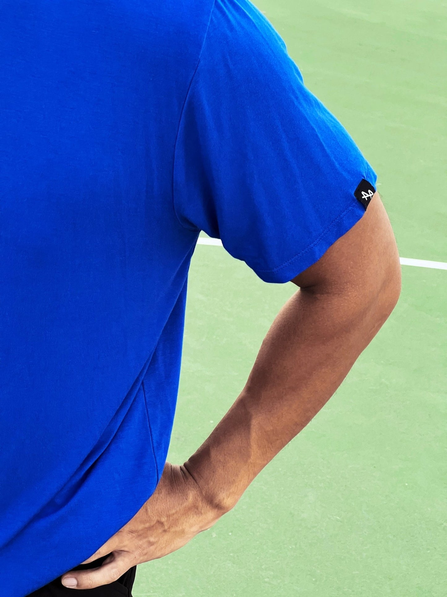 close up of ALL IN ALL blue supernatural tee left sleeve featuring sewn on black woven label with white double "A" ALL IN ALL logo against green tennis court with white court line